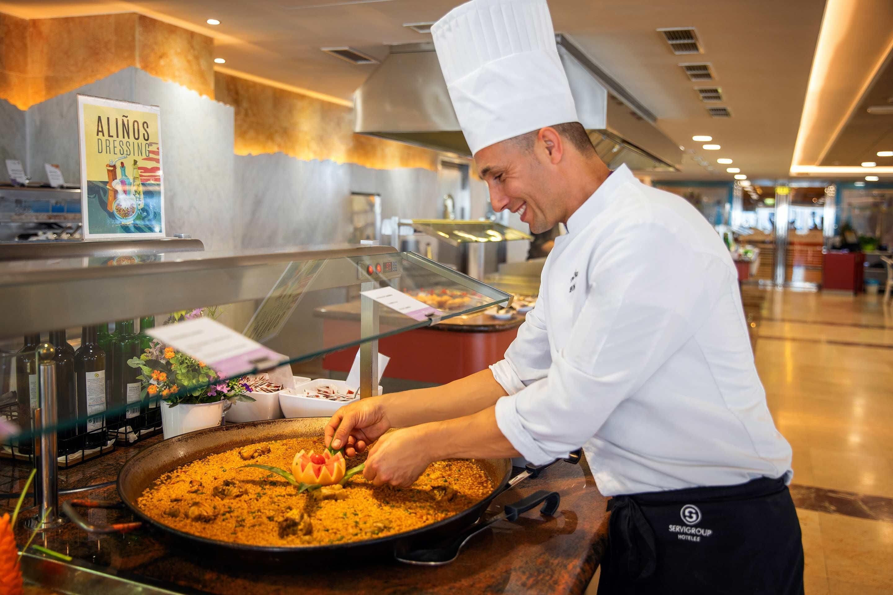 A chef preparing a dish in a professional kitchen with a poster of 'Alinos Dressing' on the wall.