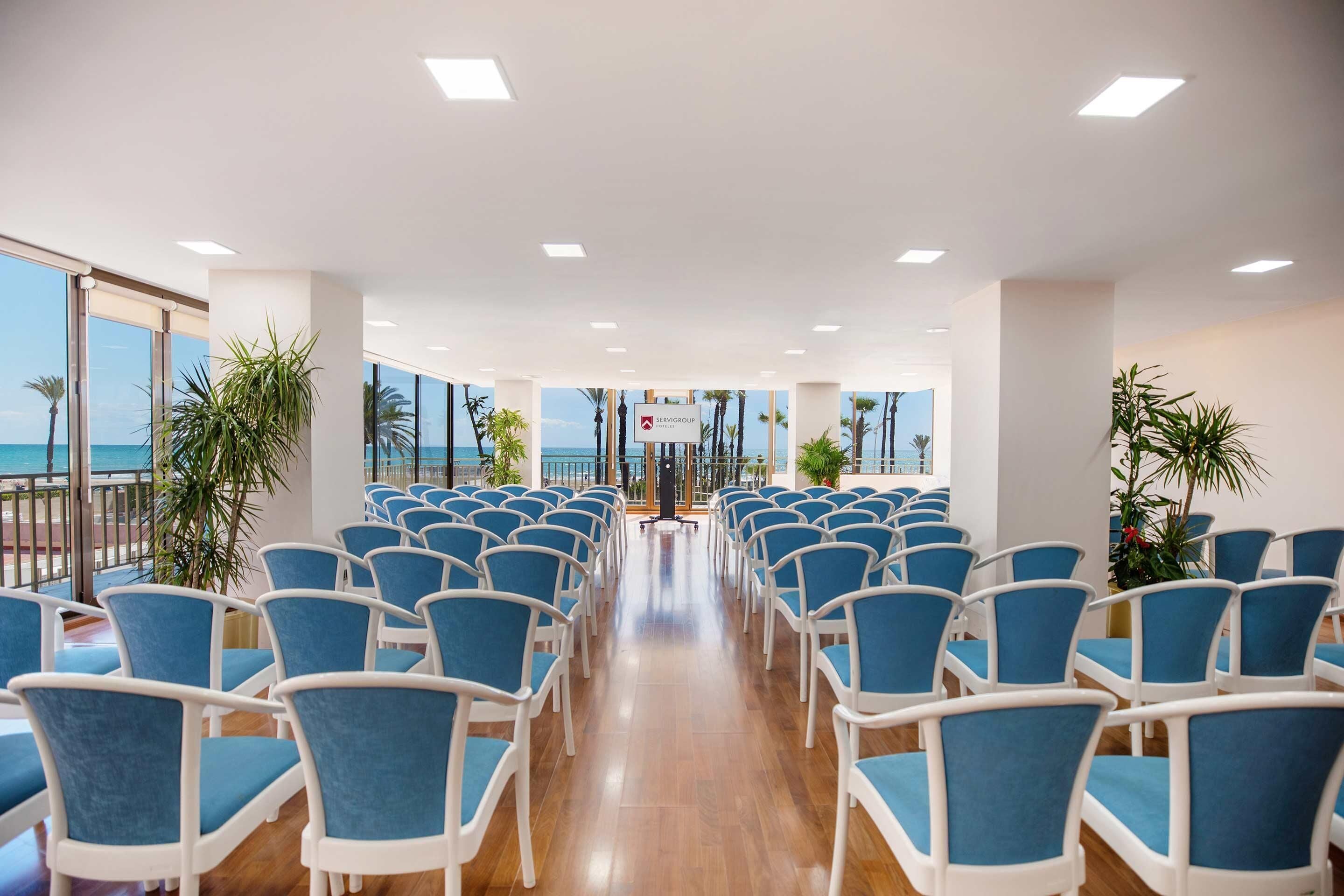 Un salón de conferencias con filas de sillas azules, un atril y vistas al océano a través de amplias ventanas.