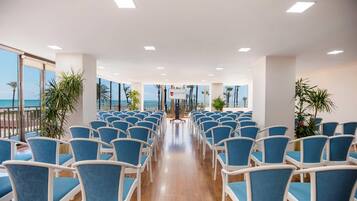 Un salón de conferencias con filas de sillas azules, un atril y vistas al océano a través de amplias ventanas.