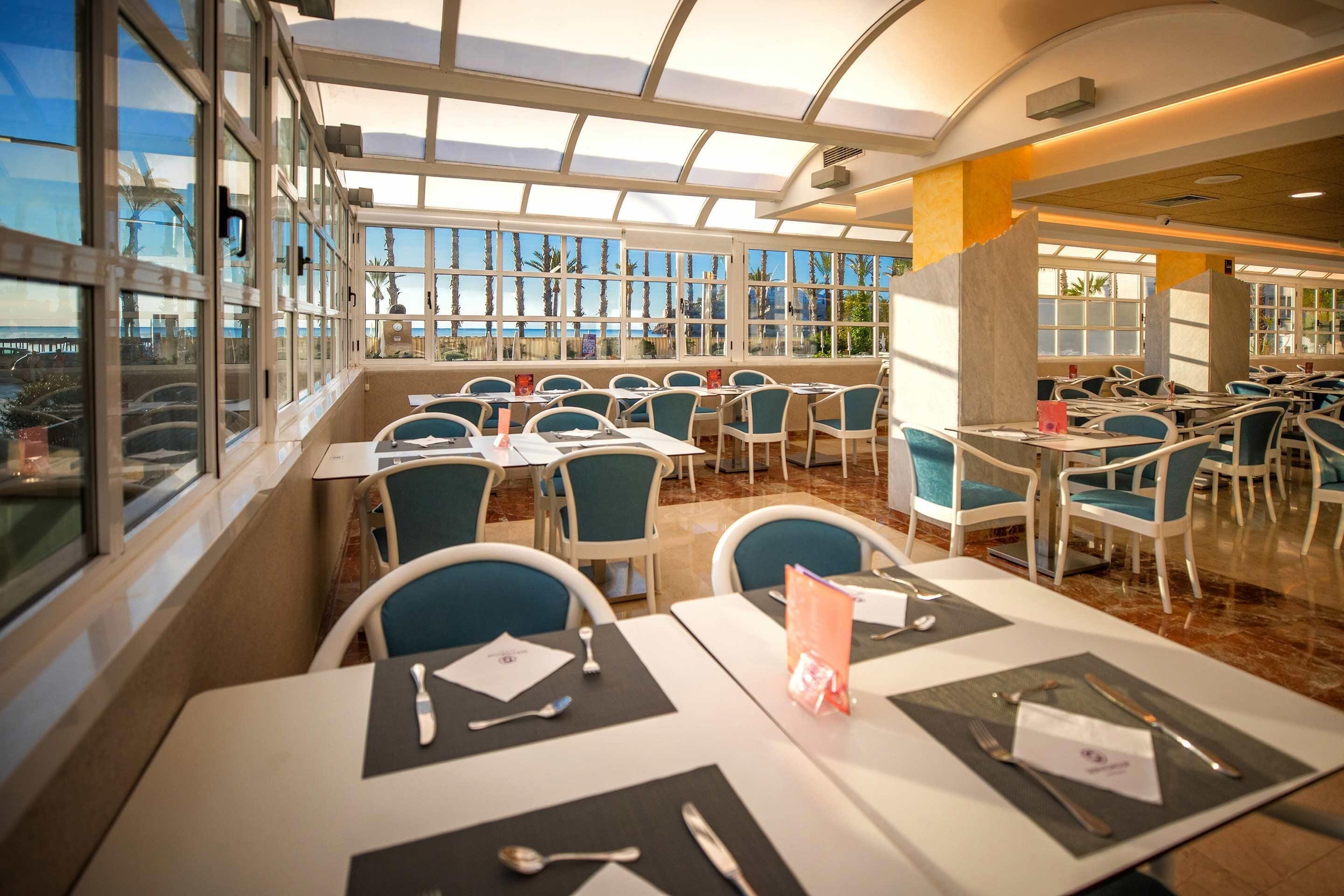 A restaurant with a terrace, tables with chairs, and a view of palm trees and the sea.