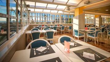 A restaurant with a terrace, tables with chairs, and a view of palm trees and the sea.