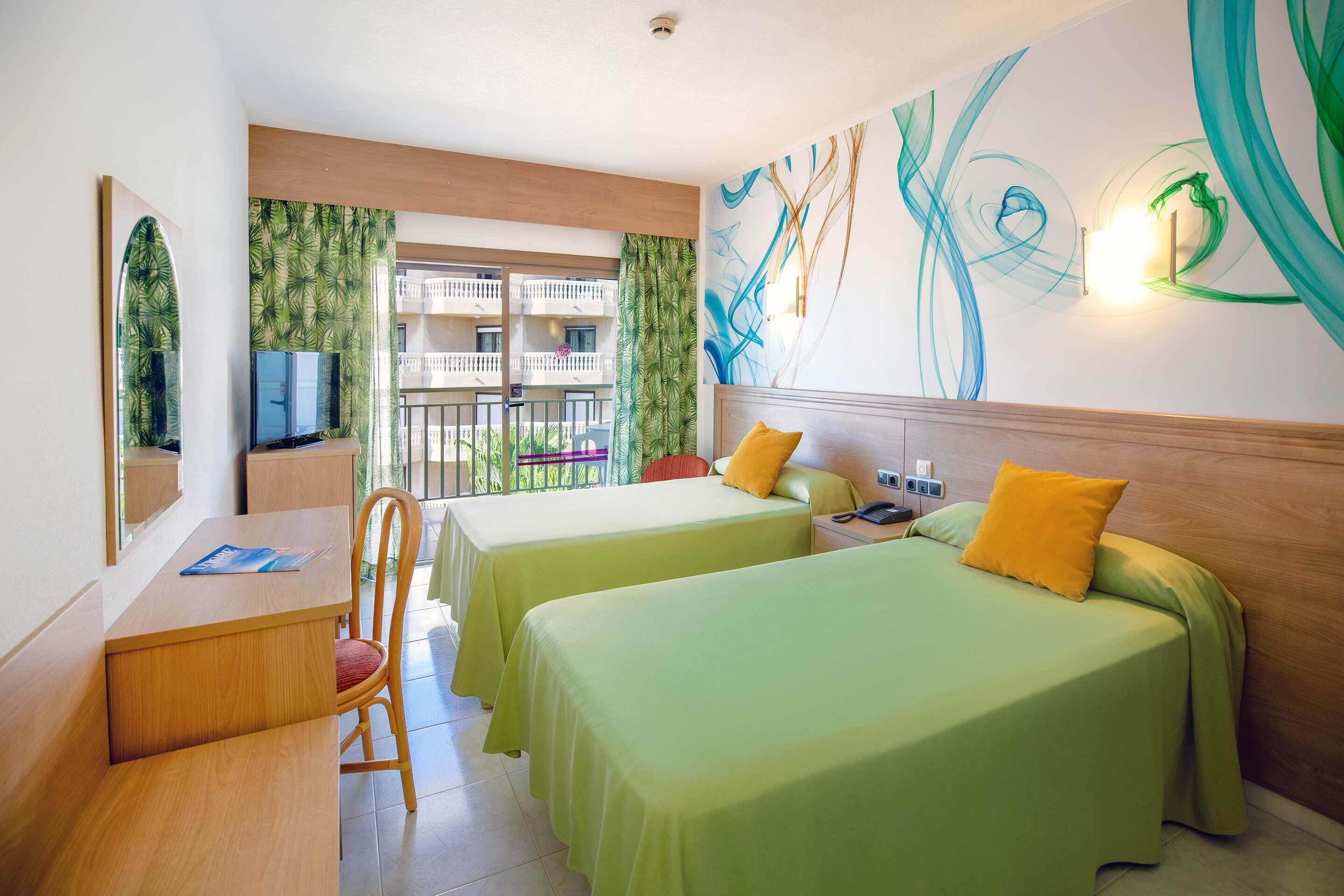A hotel room with two beds, a desk, a chair, a TV, and a balcony with a view of other buildings.