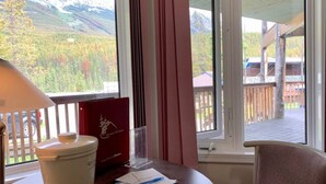 A room with a view of mountains and trees through large windows, a table with a red book and papers, a lamp, and a chair.