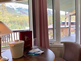 Una habitación con vistas a montañas y árboles a través de amplios ventanales, una mesa con un libro rojo y papeles, una lámpara y una silla.