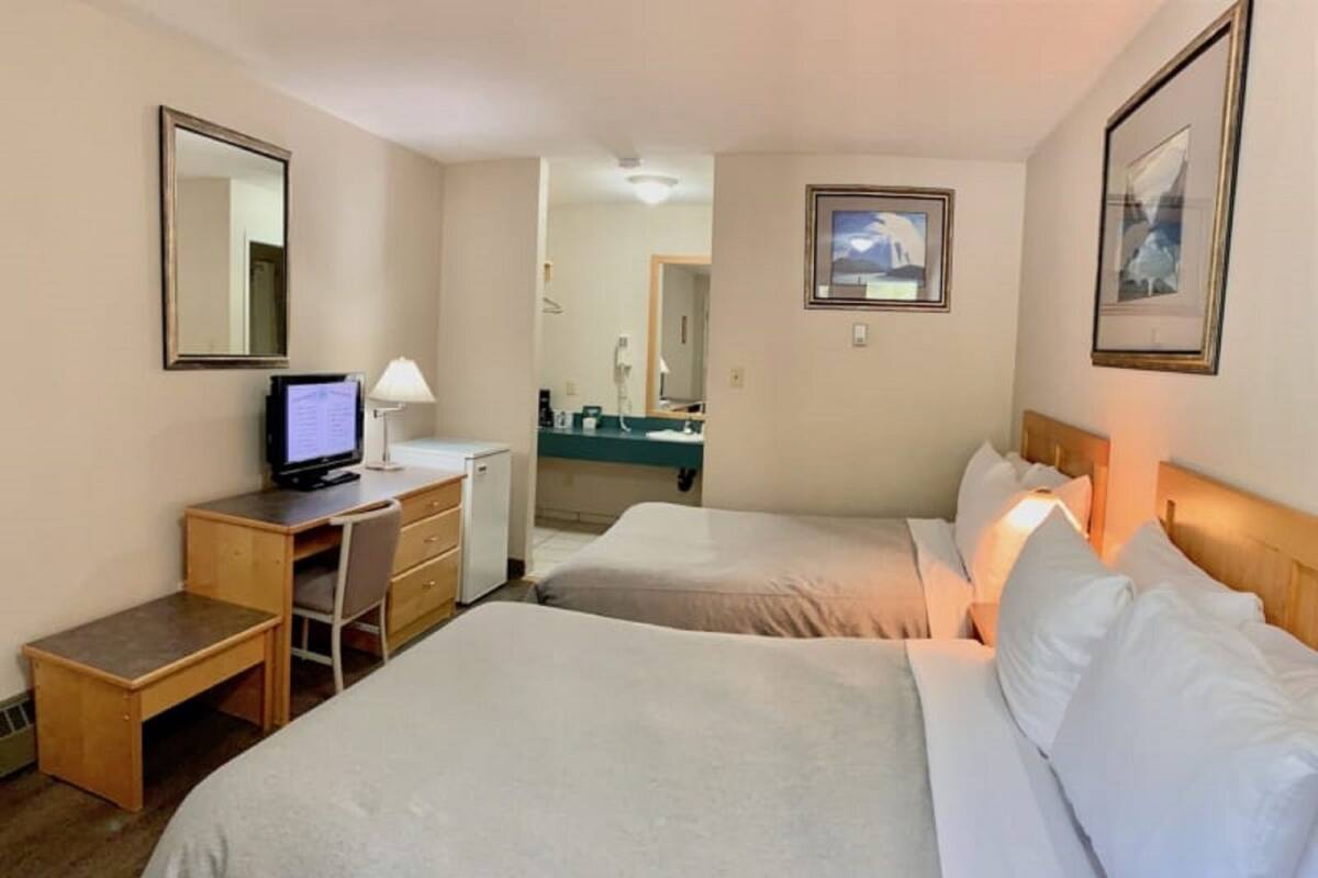 Ein Hotelzimmer mit zwei Betten, einem Schreibtisch mit Computer, einem kleinen Kühlschrank und einem im Hintergrund sichtbaren Badezimmer.
