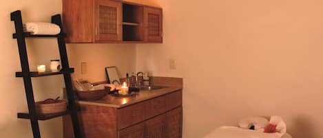 Un baño ordenado con tocador de madera, repisas con toallas, lavamanos con artículos de aseo personal y una cama con colcha blanca.