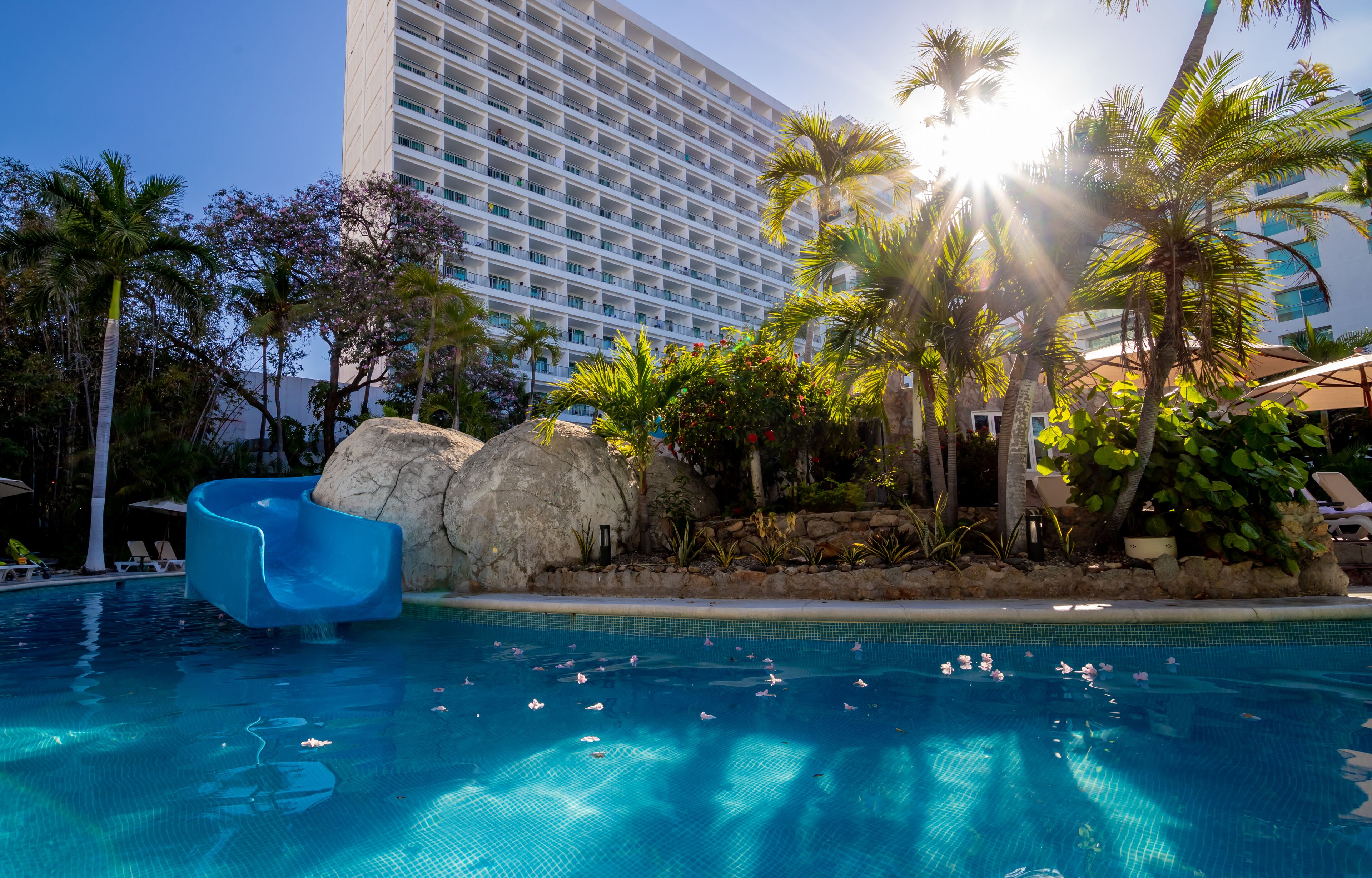 A swimming pool with a water slide, palm trees, and a large hotel building in the background.