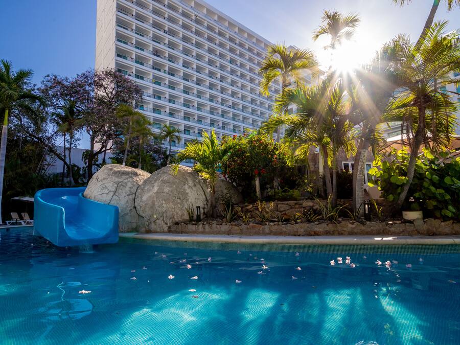 A swimming pool with a water slide, palm trees, and a large hotel building in the background.