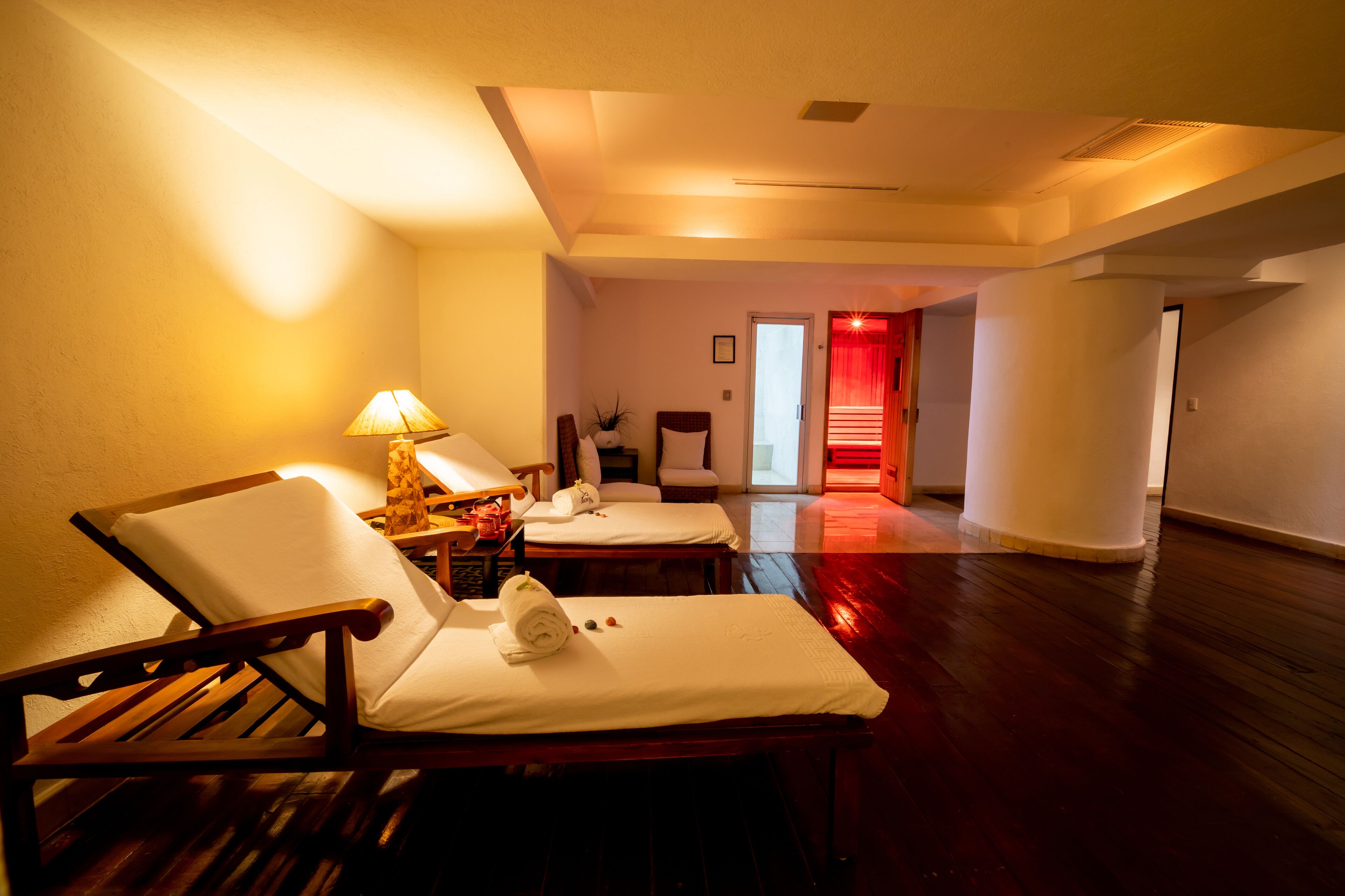 A hotel room with a wooden floor, two white cushioned chairs, a small table with a lamp, and a red-lit hallway.