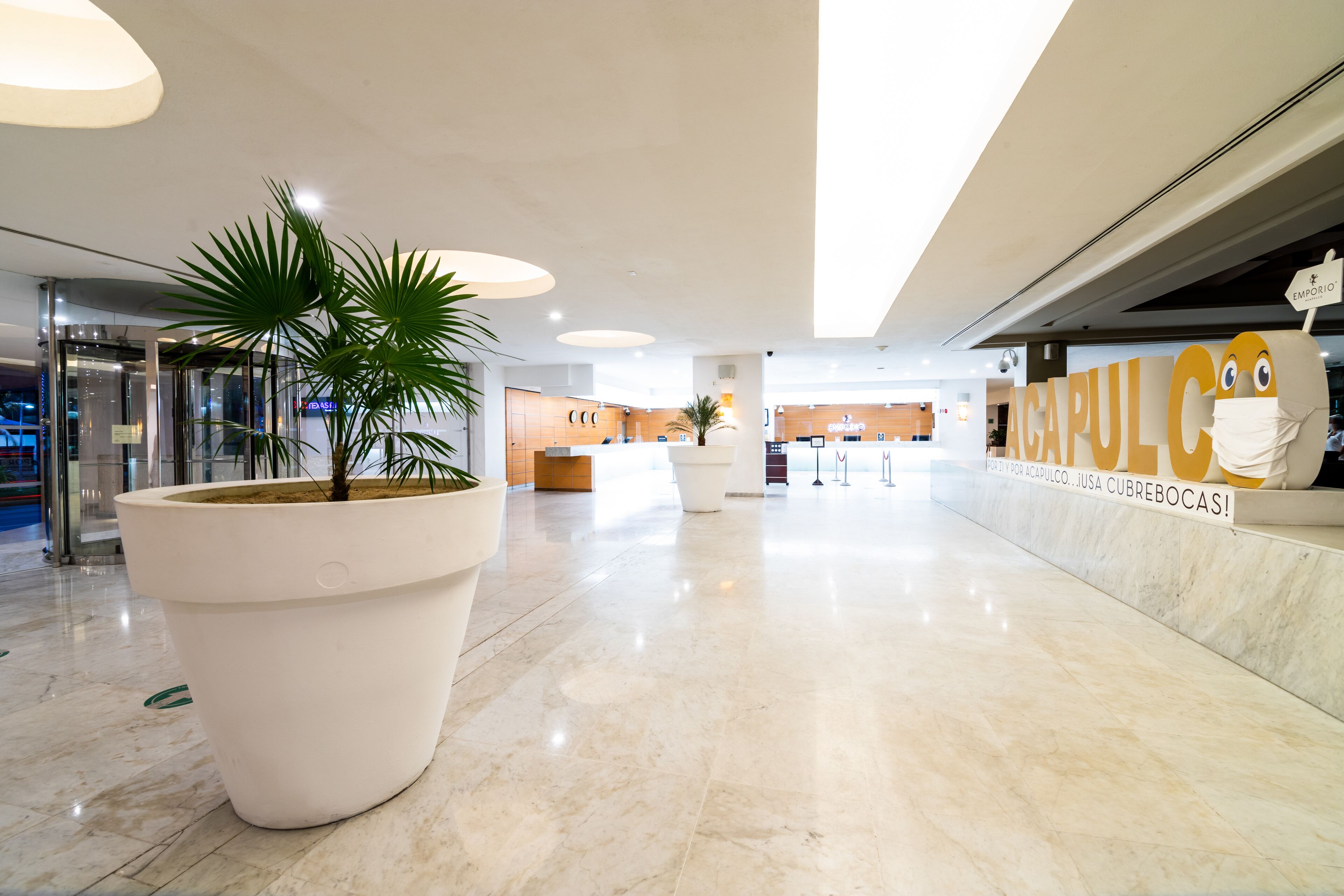 A spacious lobby with a large potted plant, a reception desk, and a sign reading "ACAPULCO".