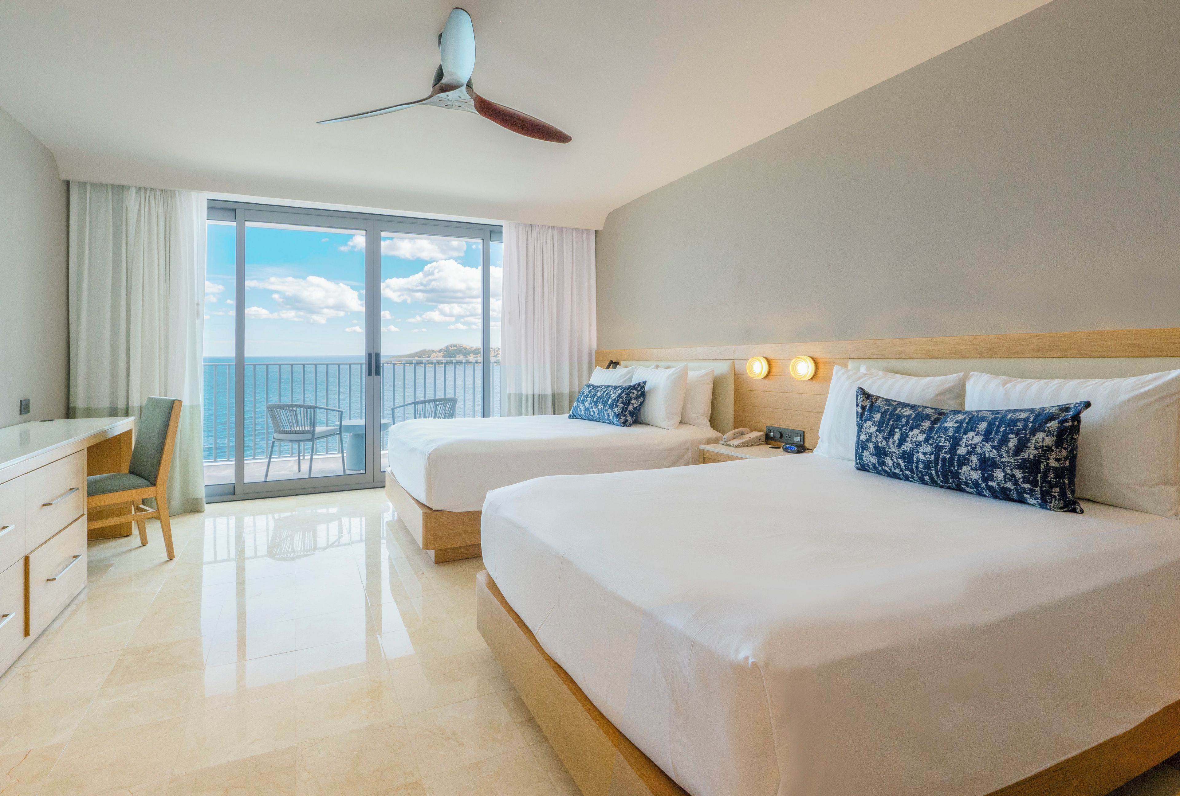 A hotel room with two beds, a desk, a chair, and a view of the sea.