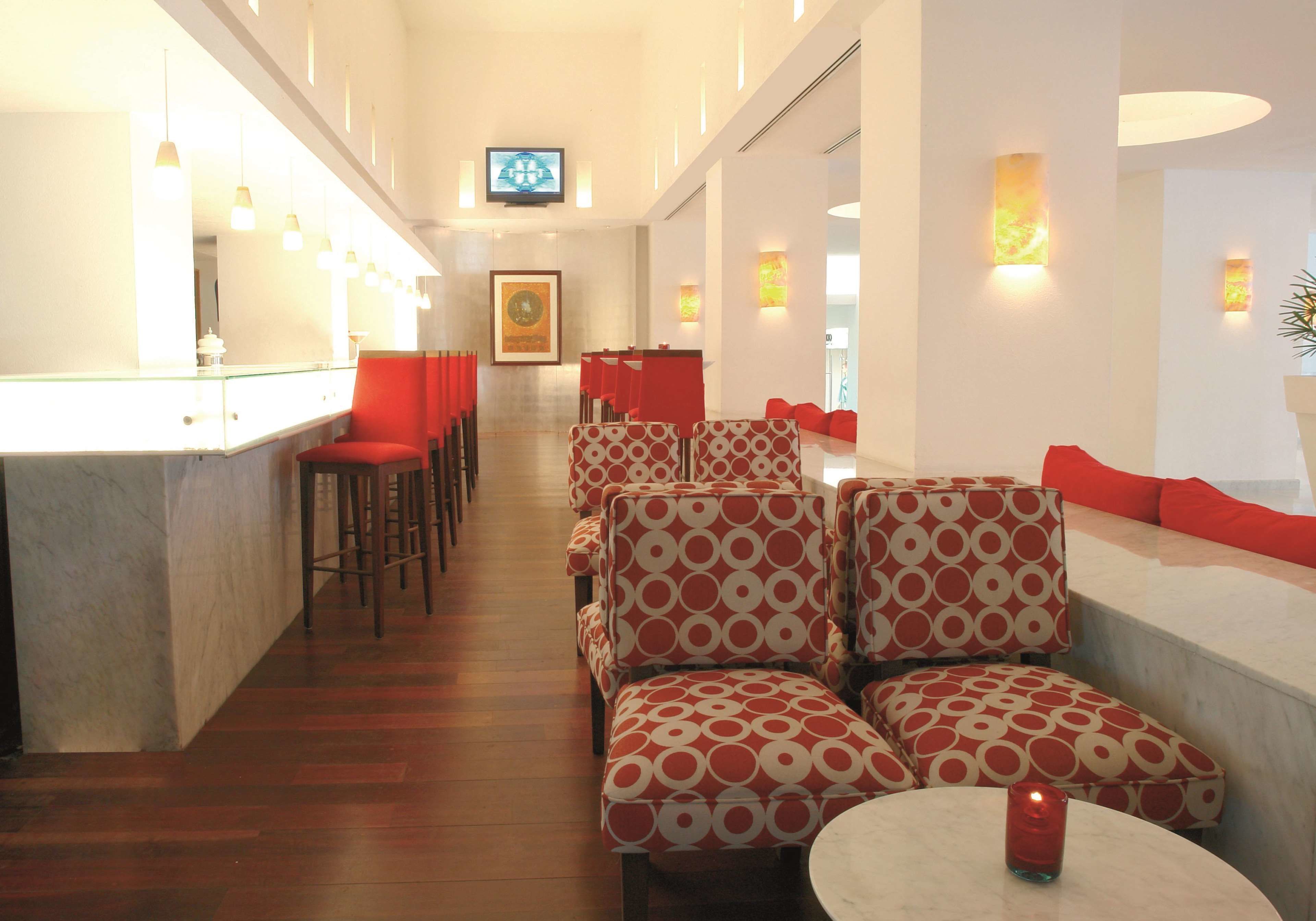 Un vestíbulo de hotel con zona de bar, asientos con cojines rojos e iluminación en la pared.