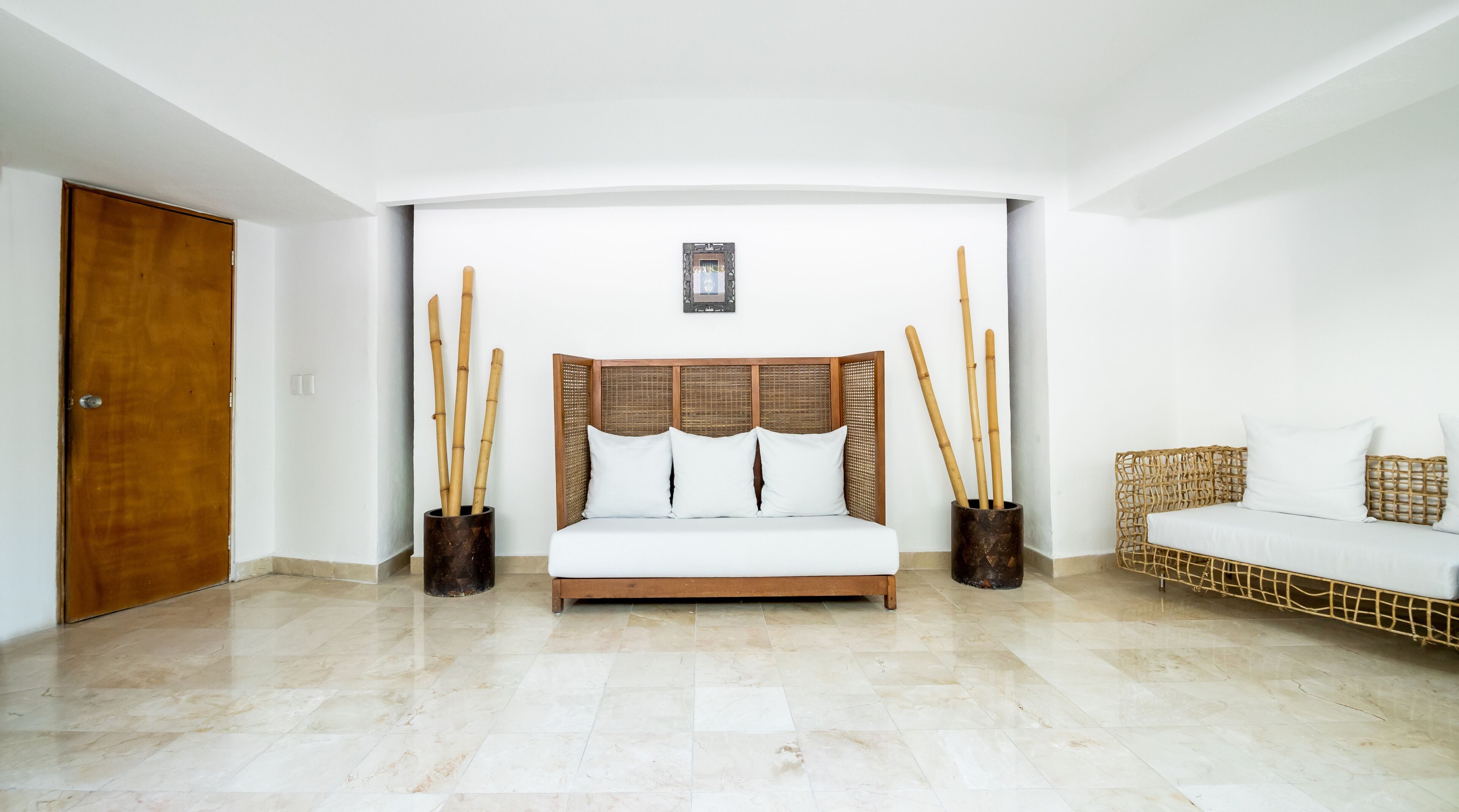 Una habitación amplia con una cama grande, dos mesitas de noche con detalles de bambú, un sofá de mimbre y una puerta de madera.