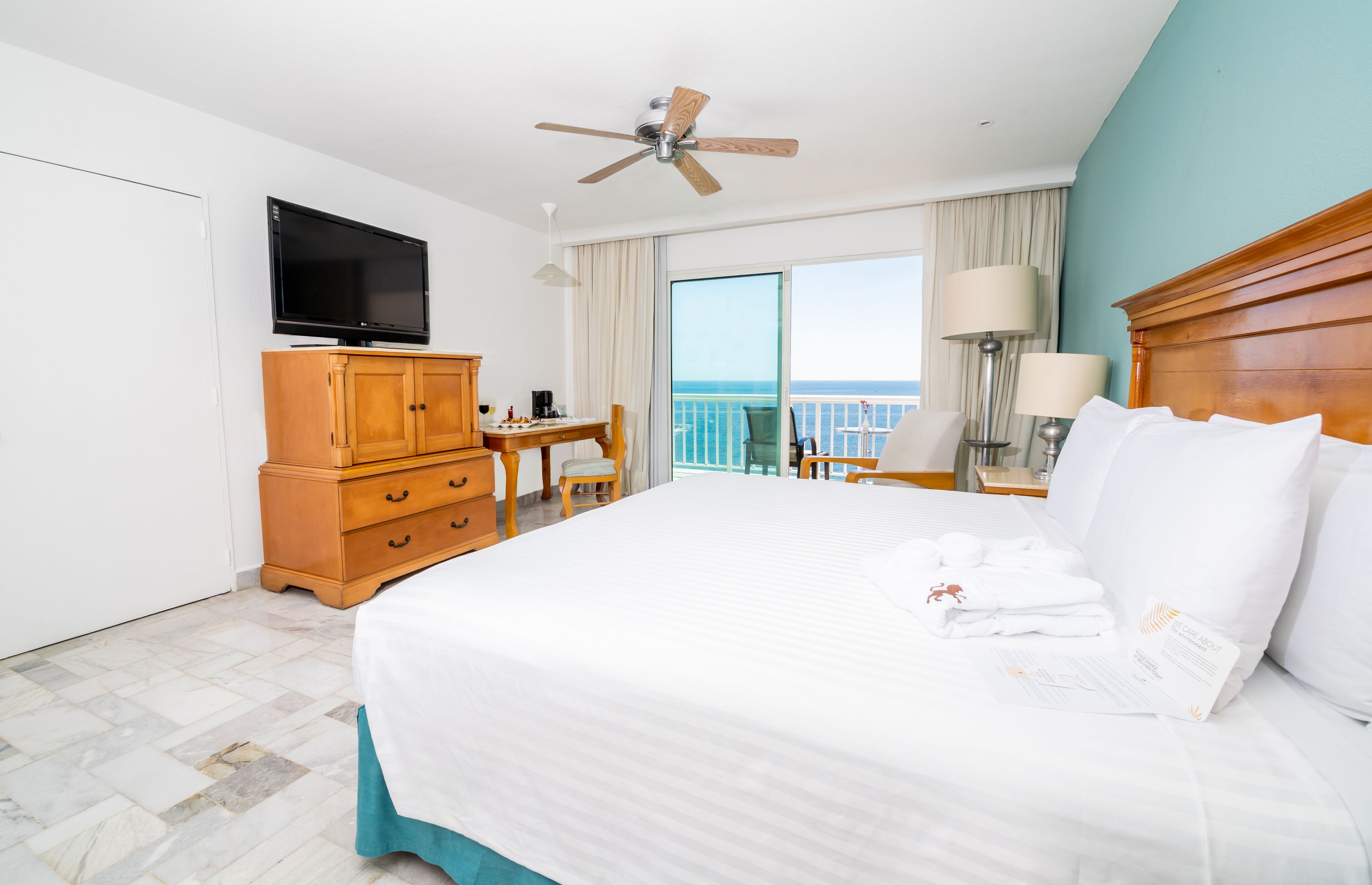 A hotel room with a bed, a TV, a wooden dresser, a desk, a chair, a balcony with a view, and a ceiling fan.
