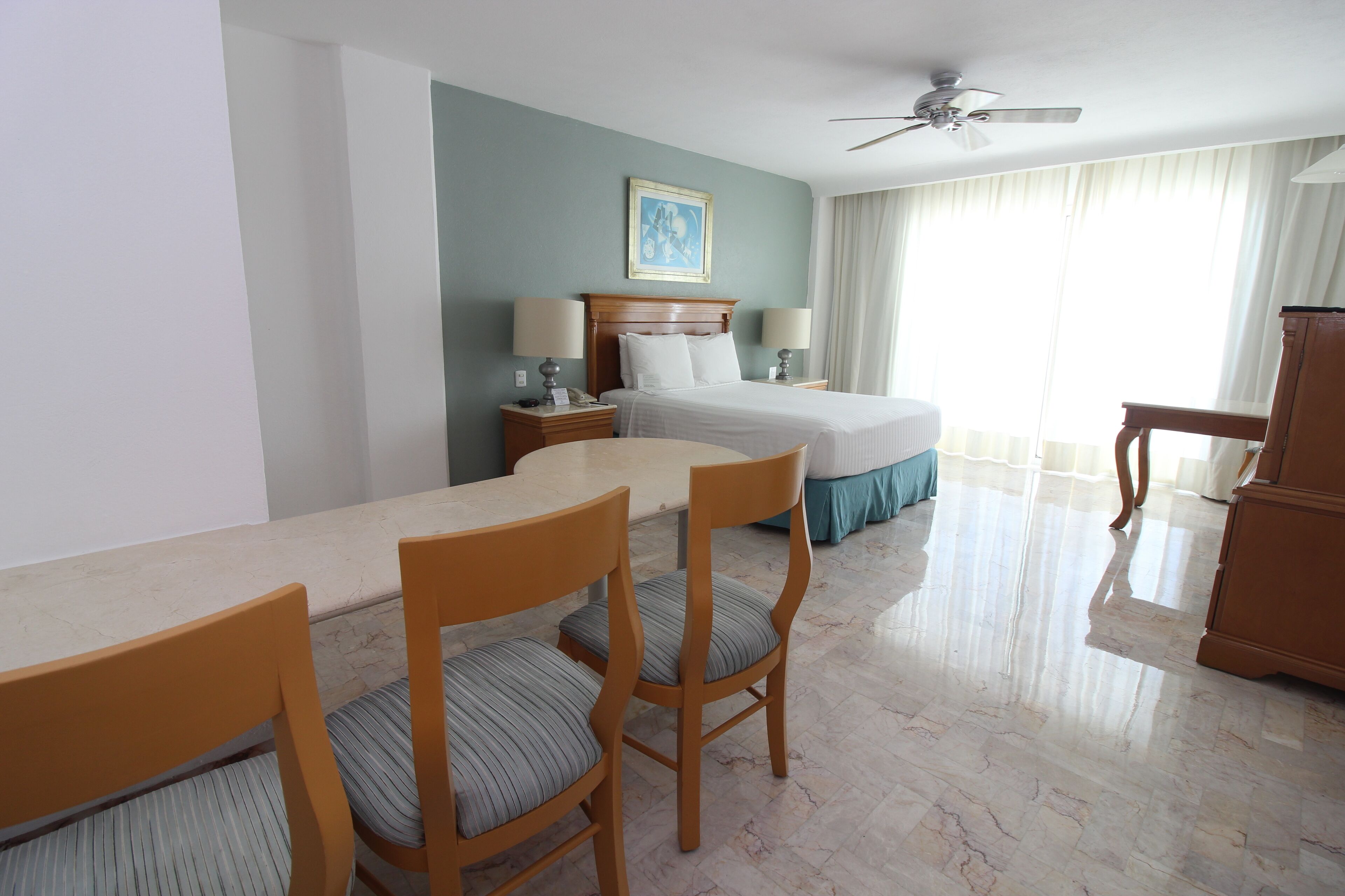 A hotel room with a bed, a desk, two chairs, a ceiling fan, and a window with curtains.