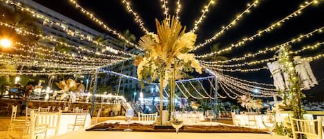 An outdoor dining area with string lights, white tables, and chairs, surrounded by a building with balconies.