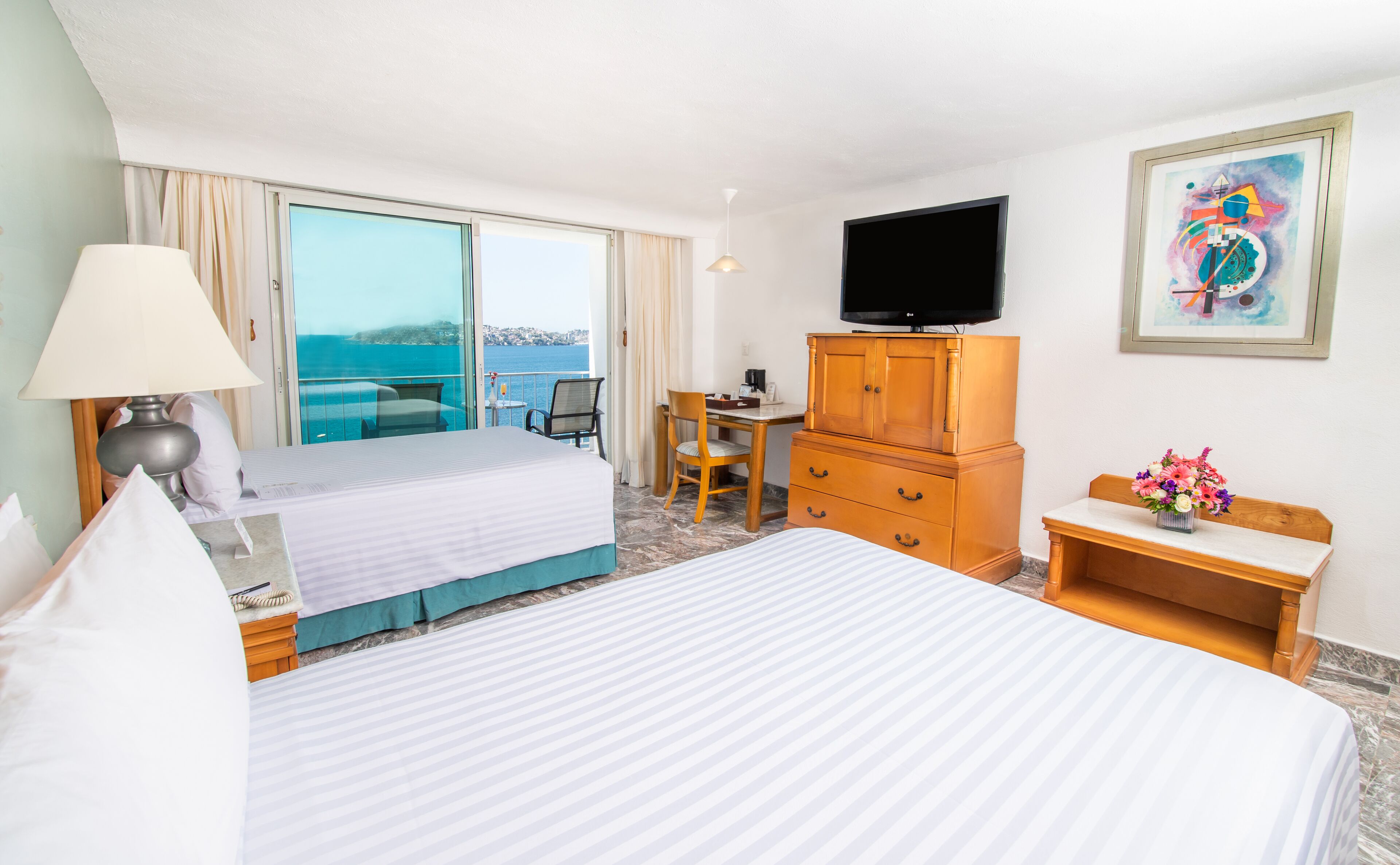 A hotel room with a bed, a desk, a chair, a television, and a view of the sea.