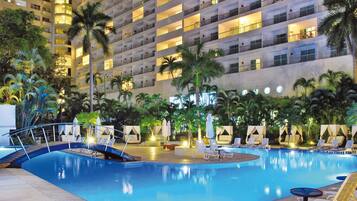 A hotel pool area with a clear blue pool, lounge chairs, palm trees, and a multi-story building in the background.