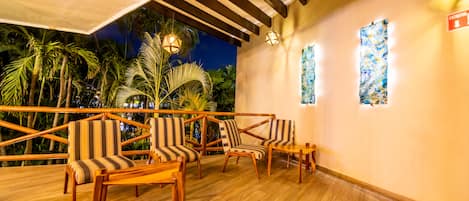 A wooden deck with striped chairs and tables, a ceiling with wooden beams, and a view of palm trees.