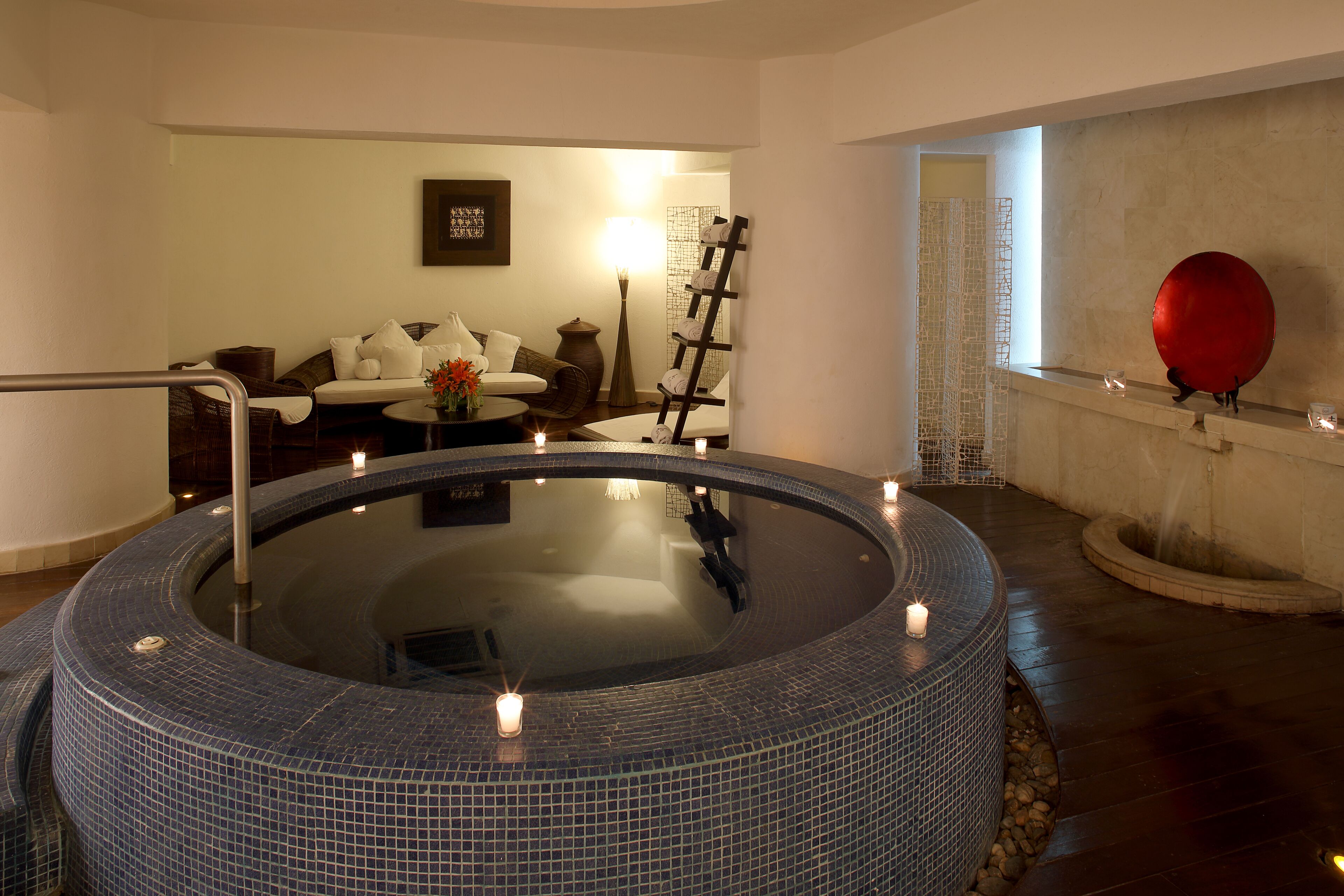 A spa area with a circular hot tub, a seating area with a sofa and armchairs, and a decorative wall with a red spherical object.