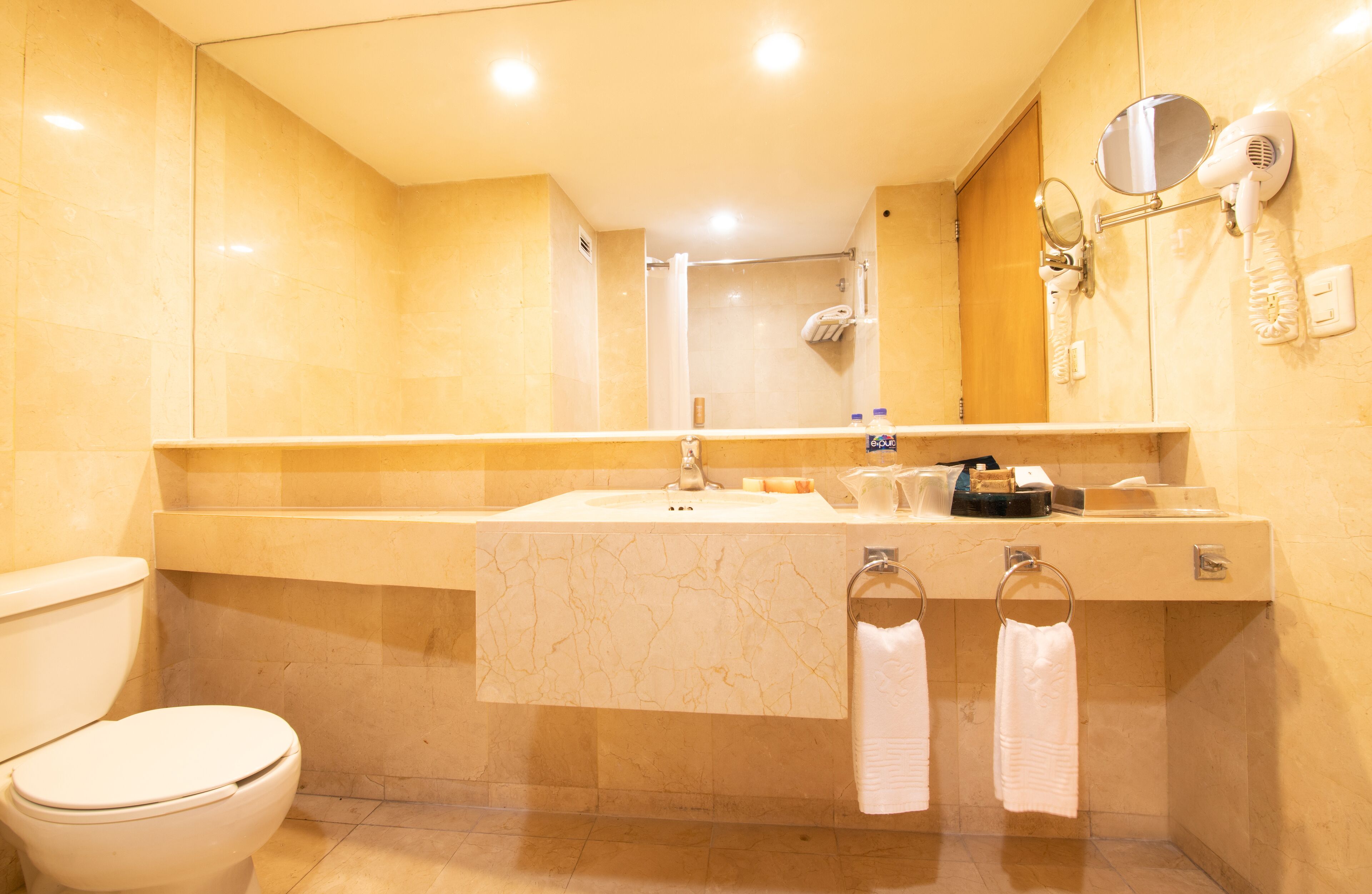 A bathroom with a toilet, sink, and mirror. There are towels hanging on hooks and a hairdryer on the wall.