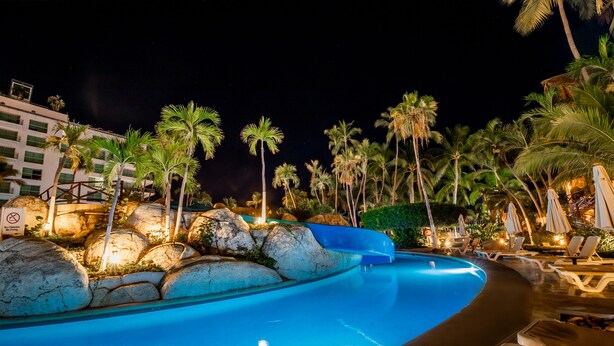 Piscina con tobogán de agua, rodeada de palmeras y sillas de descanso.