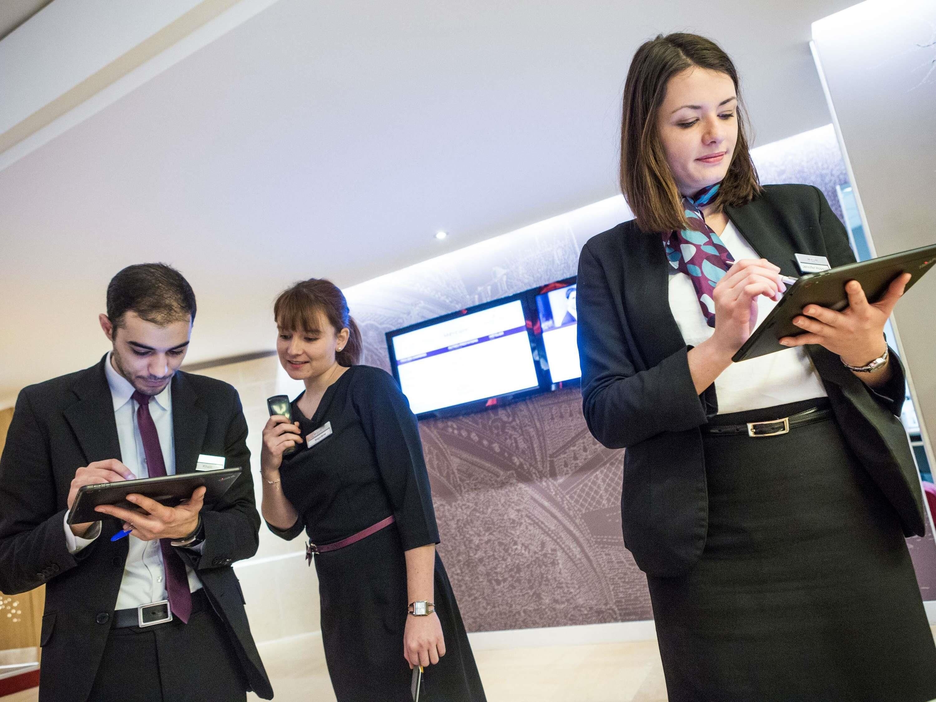 Three individuals in business attire using tablets in a modern indoor setting with a large screen displaying content.