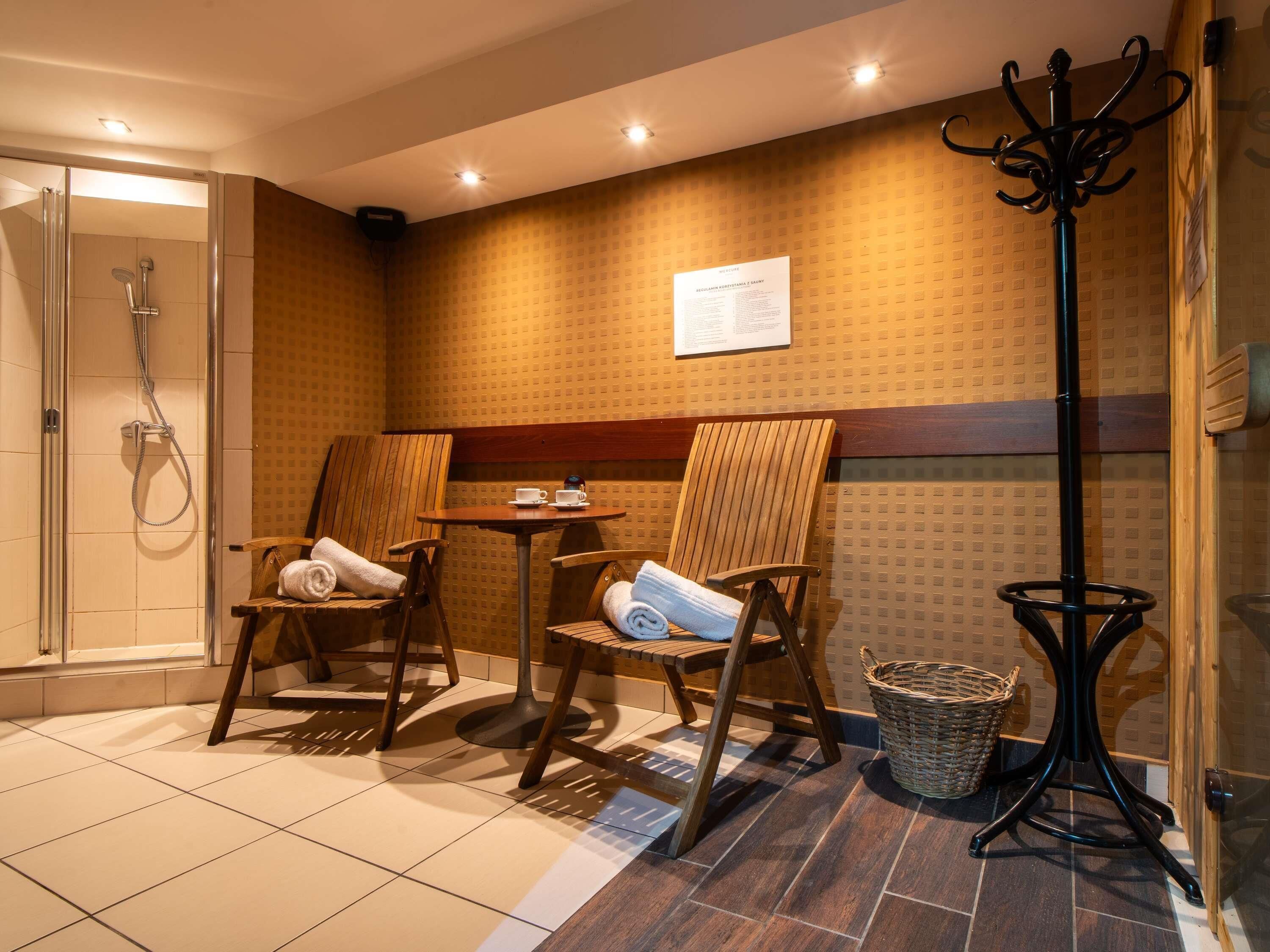 A bathroom with a shower, two wooden chairs, a small table with a cup and saucer, and a black floor lamp.