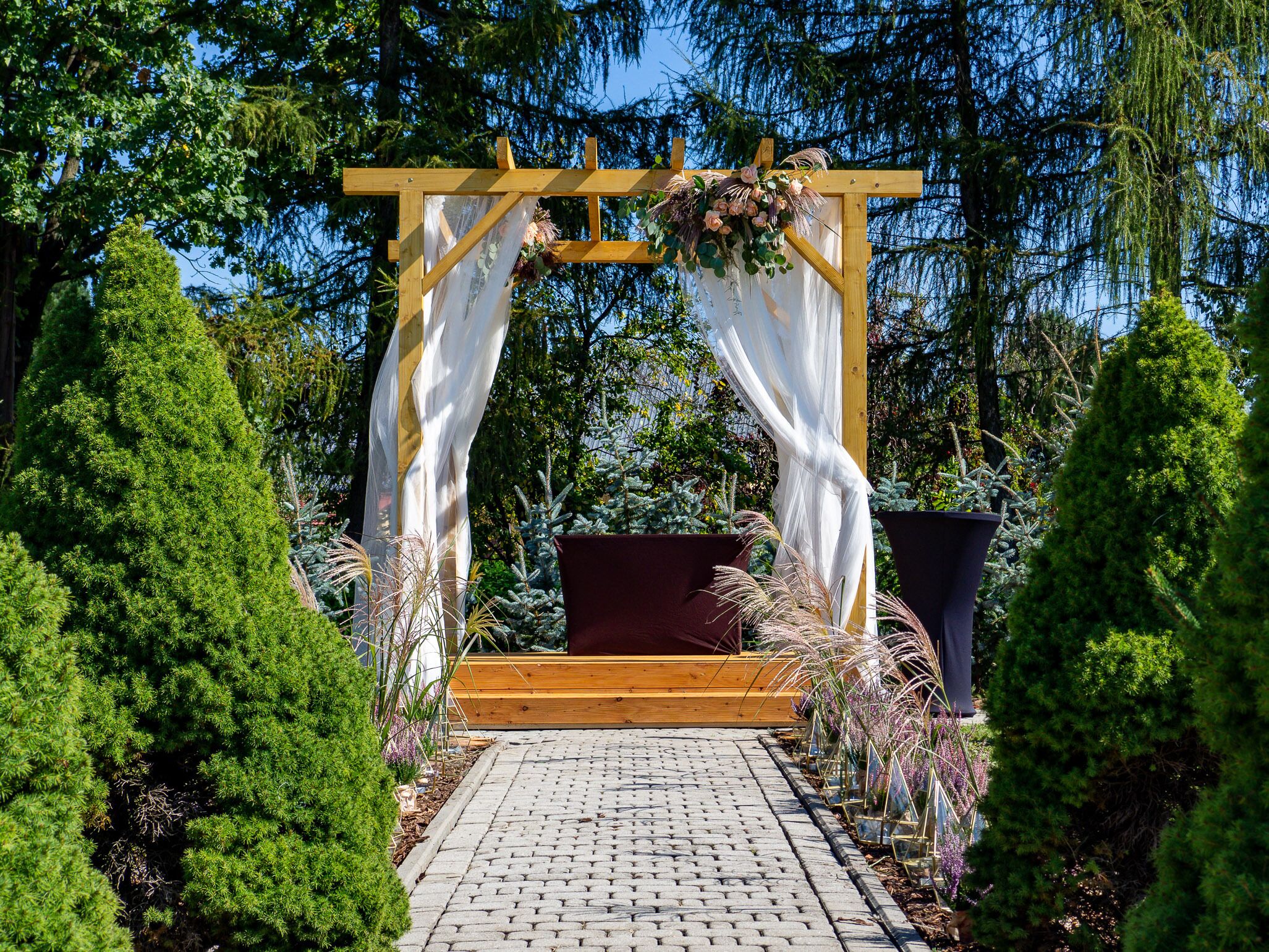 A wooden arbor with white drapes and floral decorations, surrounded by potted plants and trees.