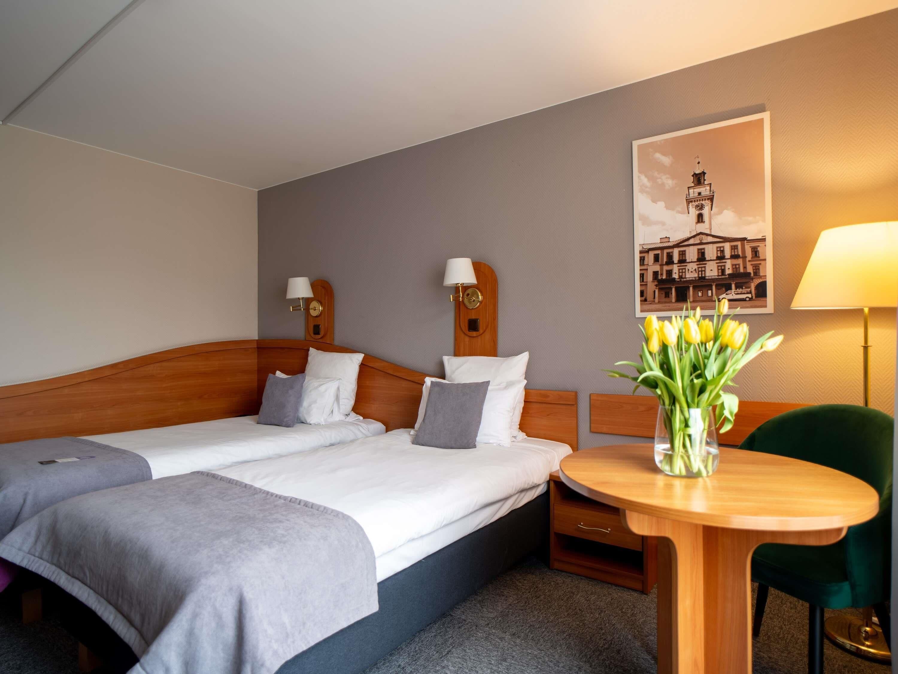 A hotel room with two beds, a round wooden table, a green chair, a lamp, and a wall-mounted picture.