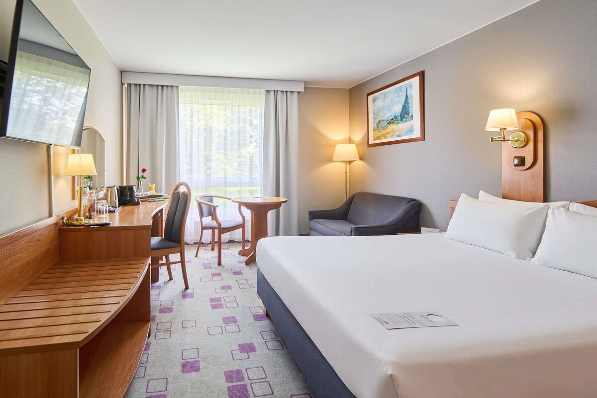 A hotel room with a bed, a desk, a chair, a small table, a TV, a lamp, and a painting on the wall.