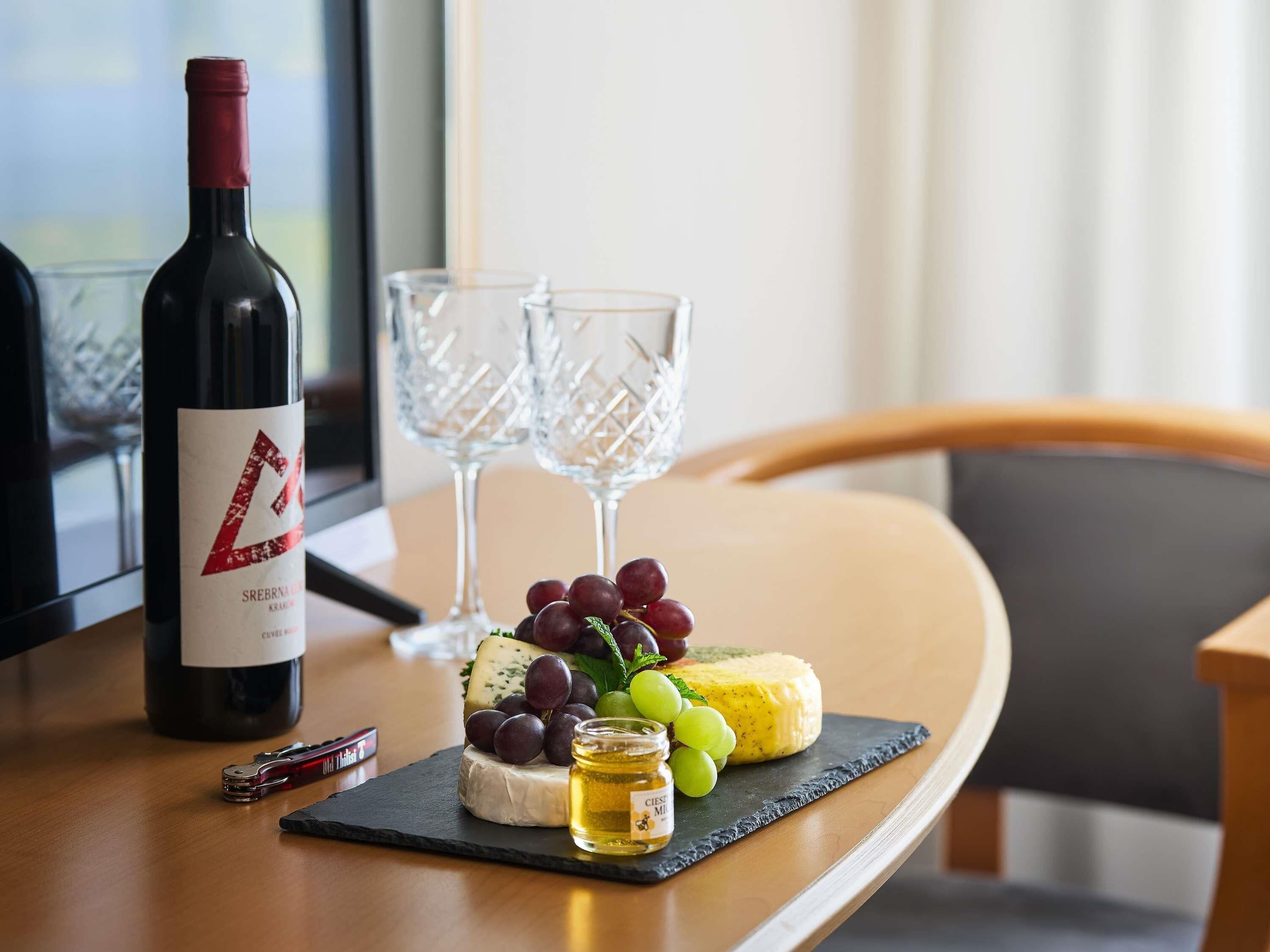 A table with a bottle of wine, two glasses, a cheese platter with grapes, and a small jar of honey.