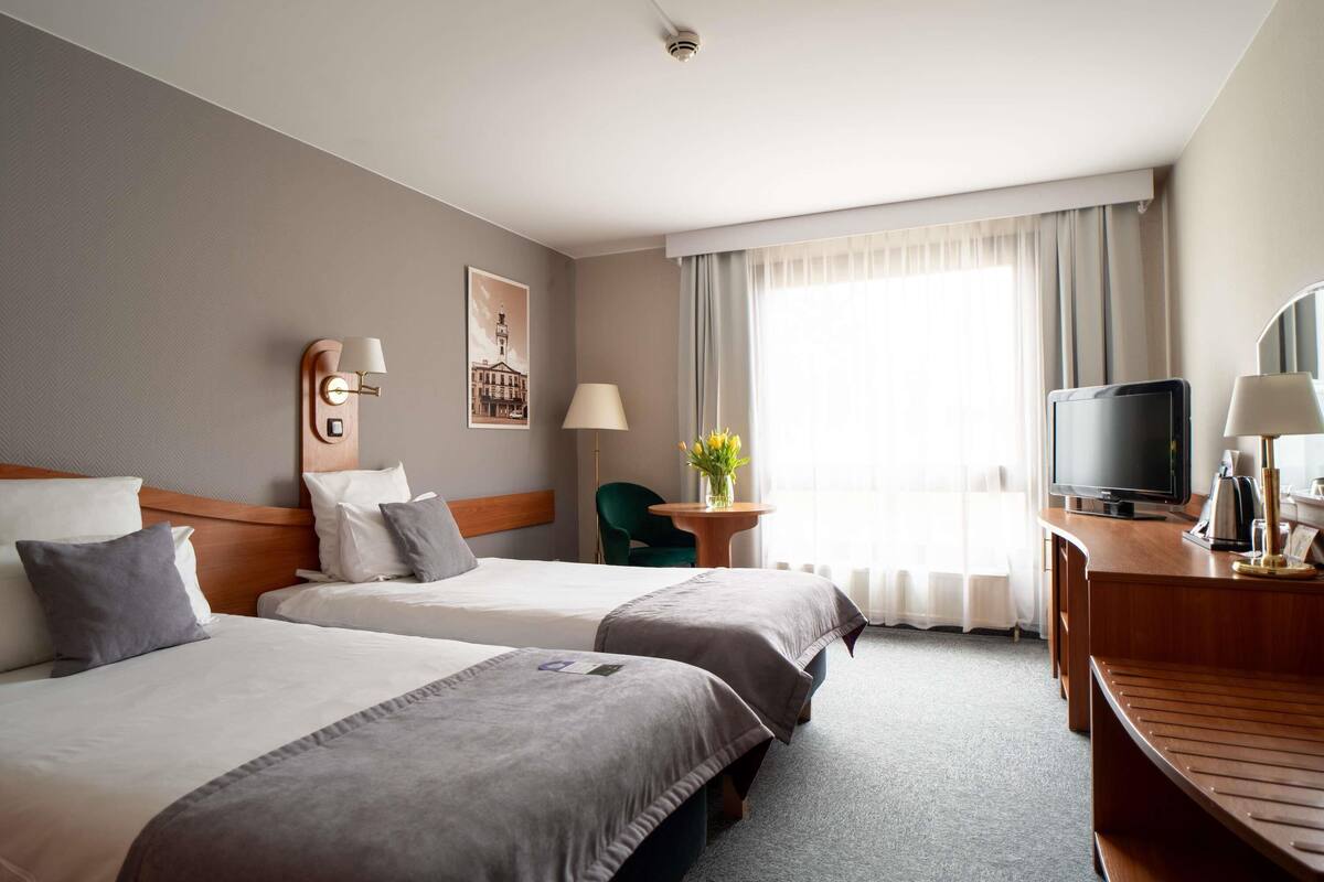 A hotel room with two beds, a desk with a television, a chair, a lamp, and a window with curtains.