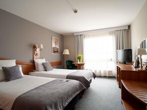A hotel room with two beds, a desk with a television, a chair, a lamp, and a window with curtains.