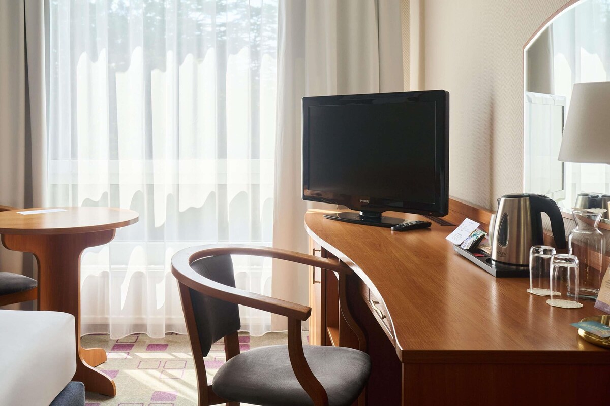 A hotel room with a wooden desk, a television, a chair, a coffee maker, and a window with sheer curtains.