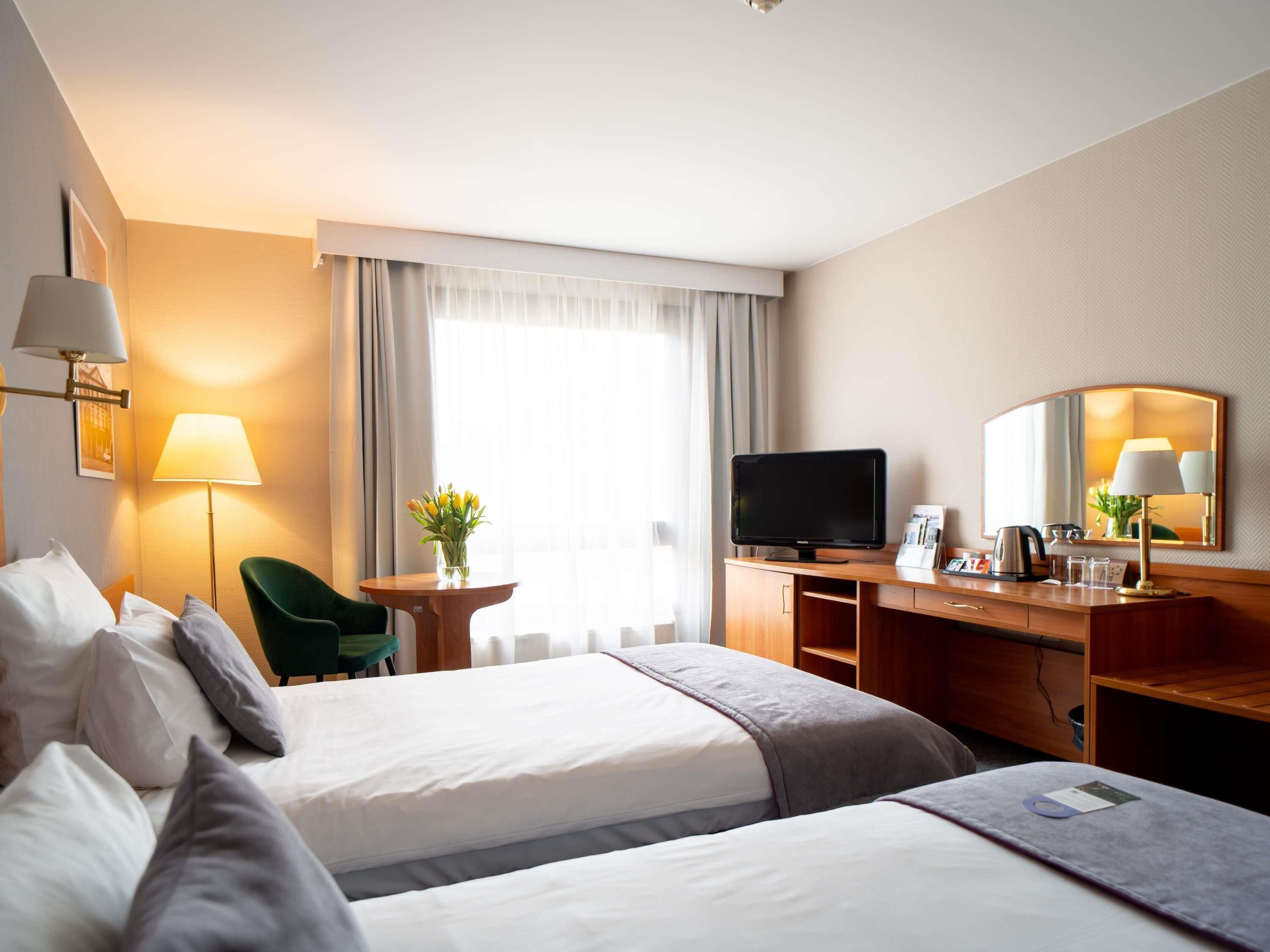 Ein Hotelzimmer mit zwei Betten, einem Schreibtisch, einem Fernseher, einem Spiegel und einem Fenster mit Vorhängen.