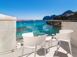 Un balcón con dos sillas blancas y una mesa, con vistas al mar azul y despejado, con barcos y terreno rocoso.