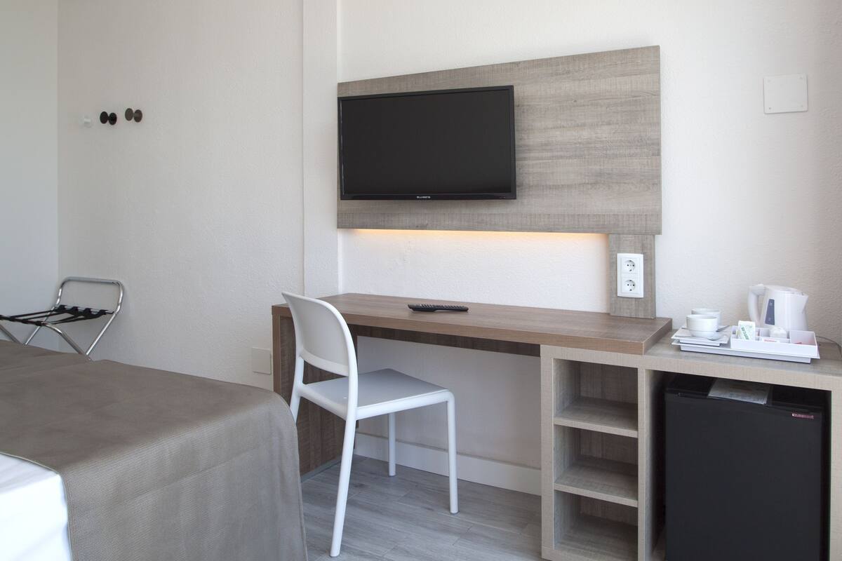 A compact hotel room with a wooden desk, a white chair, a flat-screen TV mounted above the desk, a small refrigerator, and a kettle on the desk.