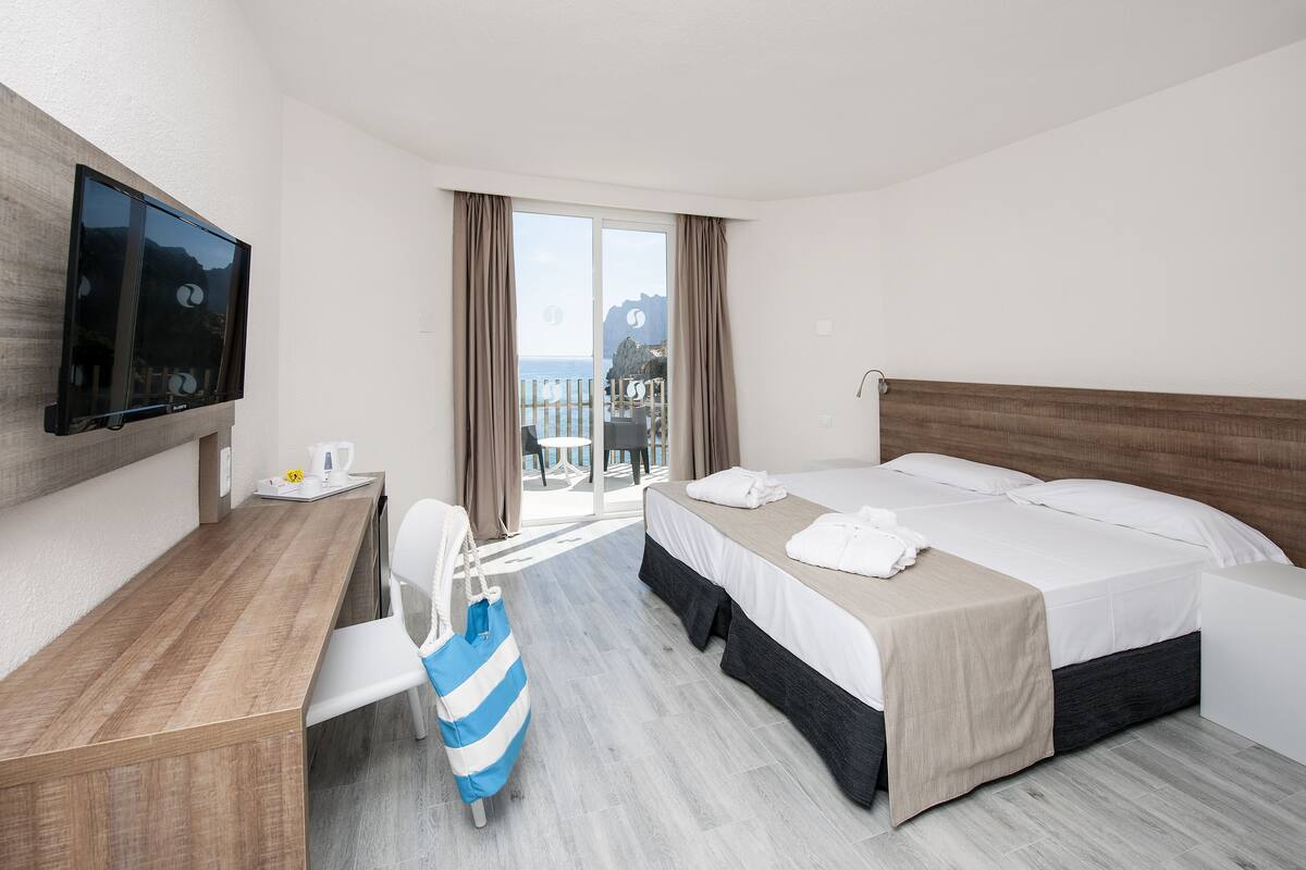 A hotel room with a bed, a desk, a TV, and a view of the sea.