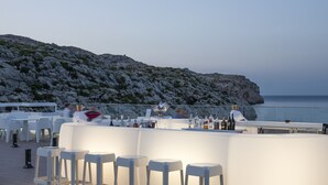 A bar area with white stools and a bar counter, overlooking a rocky coastline.