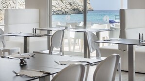 A restaurant with white chairs and tables, large windows with a view of the sea and a rocky cliff.