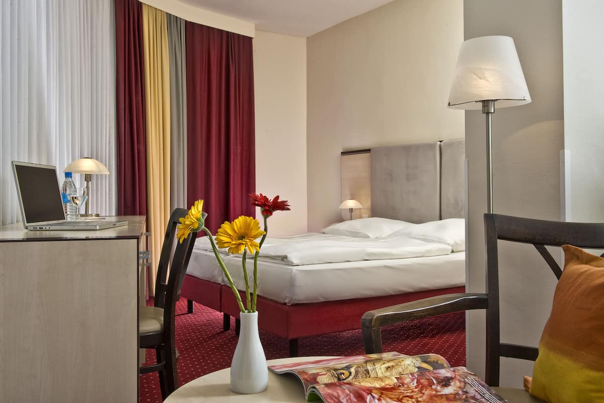 Ein Hotelzimmer mit einem Bett, einem Schreibtisch mit Laptop, einer Blumenvase und einer Zeitschrift auf dem Tisch.