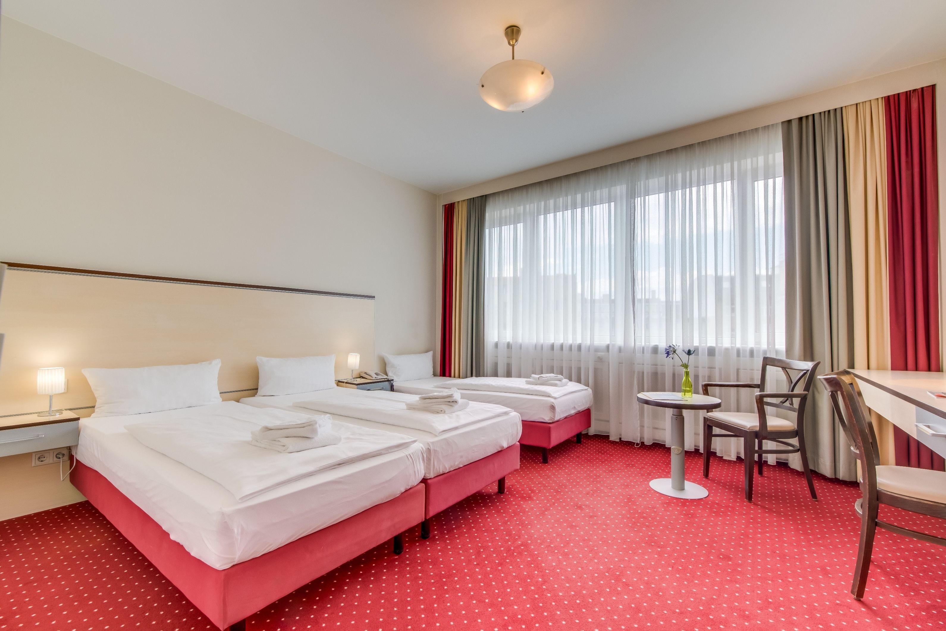 A hotel room with two beds, a desk, a chair, a small table, and a window with curtains.