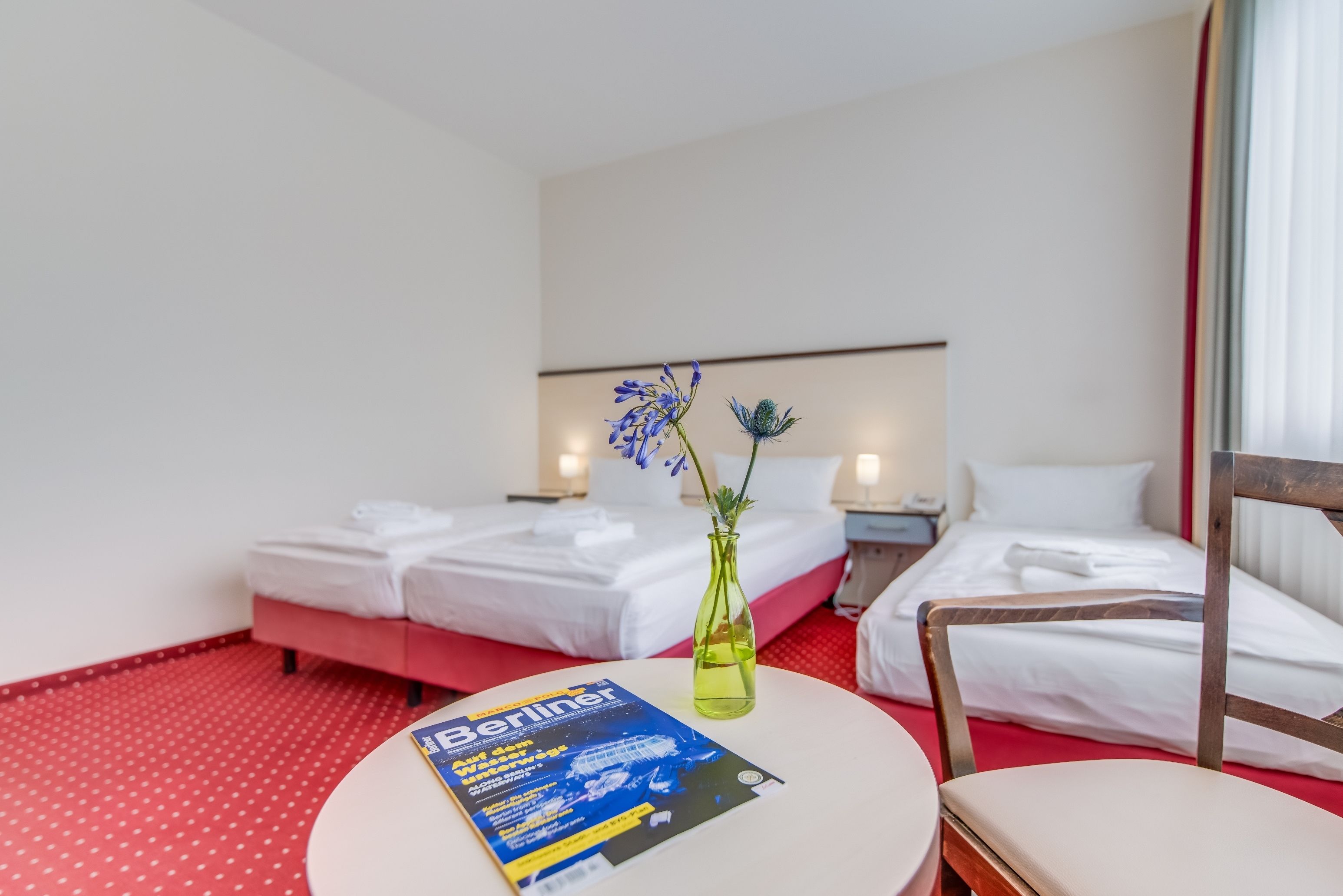 A hotel room with two beds, a small table with a magazine, and a vase with flowers.