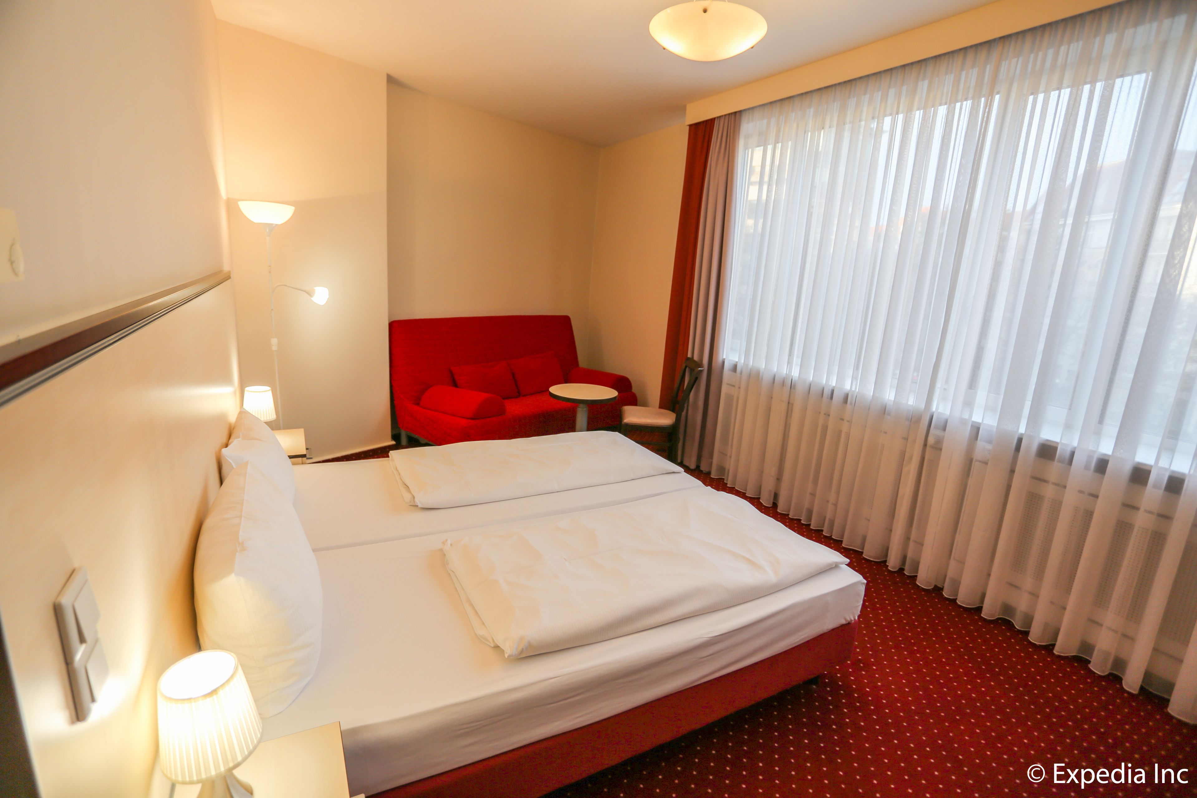 A hotel room with a bed, a red sofa, a small round table, and a window with sheer curtains.