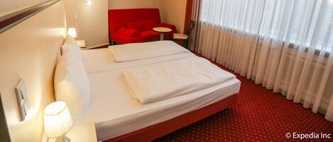 A hotel room with a bed, a red sofa, a small round table, and a window with sheer curtains.