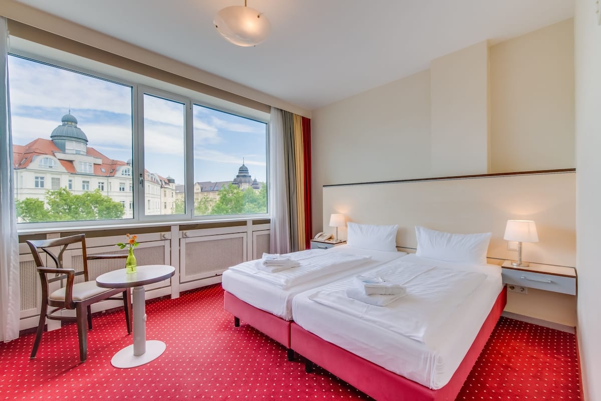 Ein Hotelzimmer mit zwei Betten, einem kleinen Tisch mit einer Vase und Blick auf Gebäude durch große Fenster.