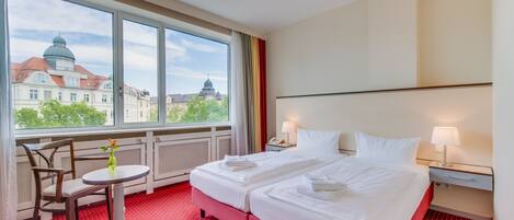 Habitación de hotel con dos camas, una mesita con un florero, una silla y ventana con vista a la ciudad.