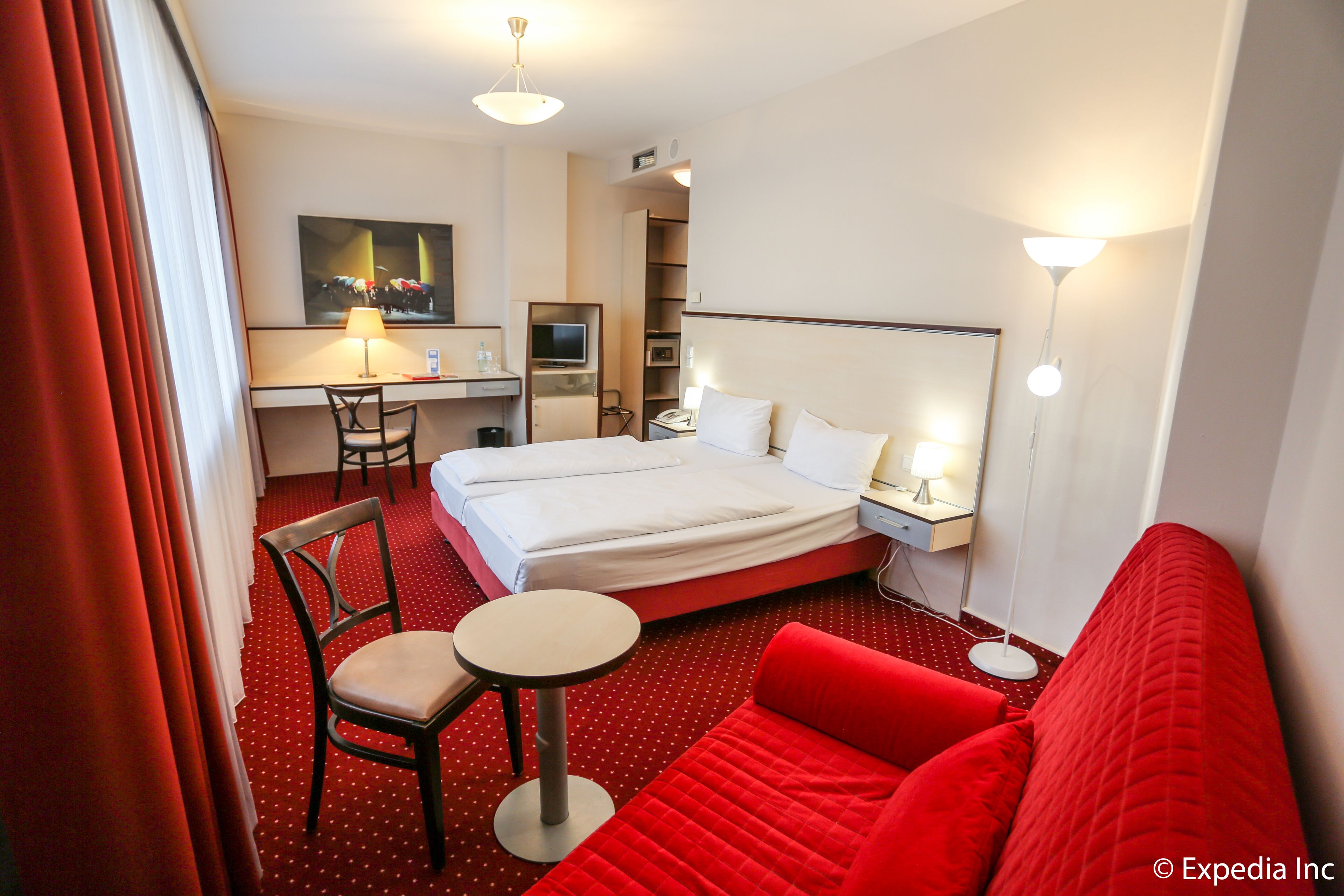Habitación de hotel con cama, sofá rojo, escritorio con silla, televisión y lámpara.