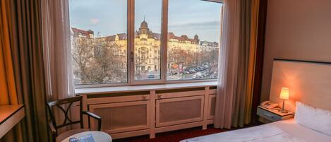 Ein Hotelzimmer mit großem Fenster und Blick auf historische Gebäude. Es gibt ein Bett, einen Nachttisch mit Lampe und einen Sessel.